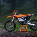 KTM Freeride E 2027 laranja, vista frontal 3/4 em trilha de terra com suspensão elevada e rodas 21/18