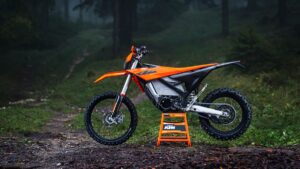 KTM Freeride E 2027: O que a marca ainda esconde KTM Freeride E 2027 laranja, vista frontal 3/4 em trilha de terra com suspensão elevada e rodas 21/18