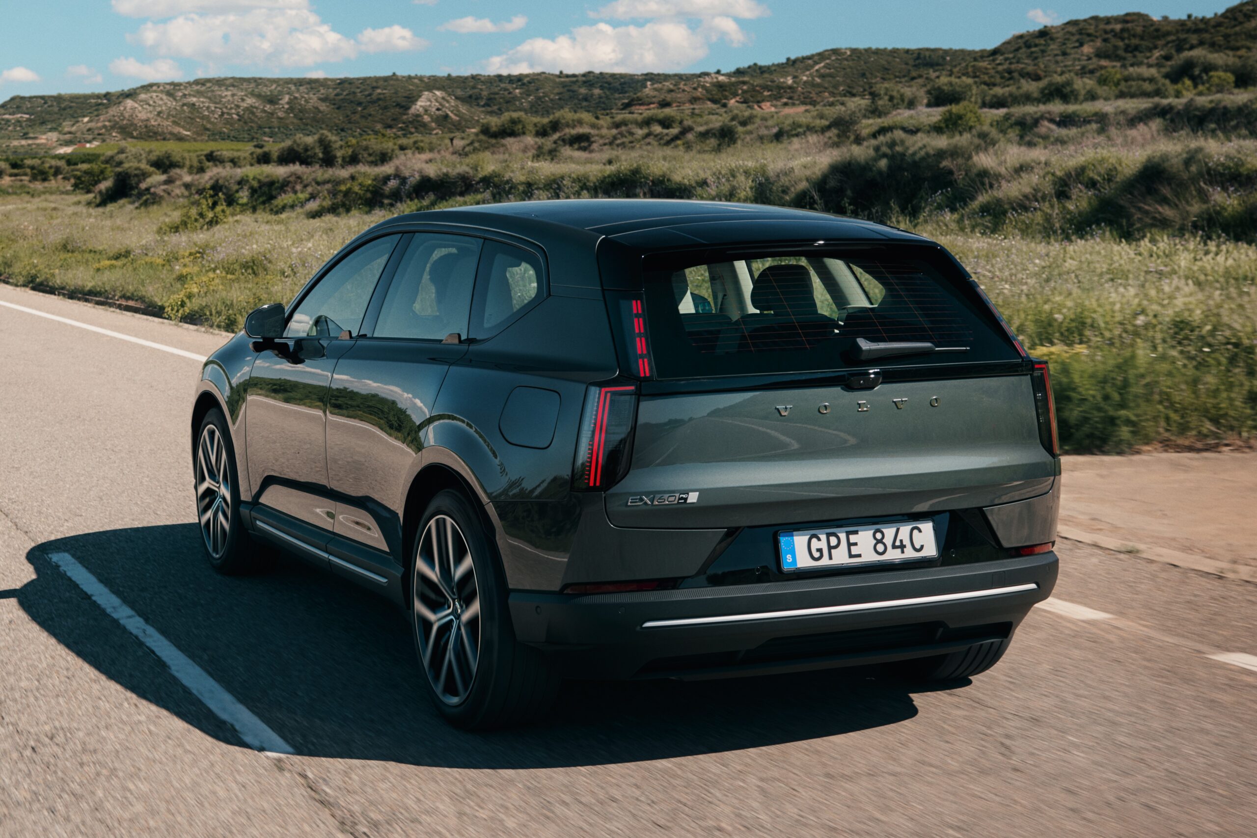 Volvo EX60 com Gemini não livra multa pelo CTB SUV Volvo EX30 cinza-escuro, visto de trás, em uma estrada asfaltada com faixa branca. Ao fundo, colinas verdes e céu azul com nuvens esparsas