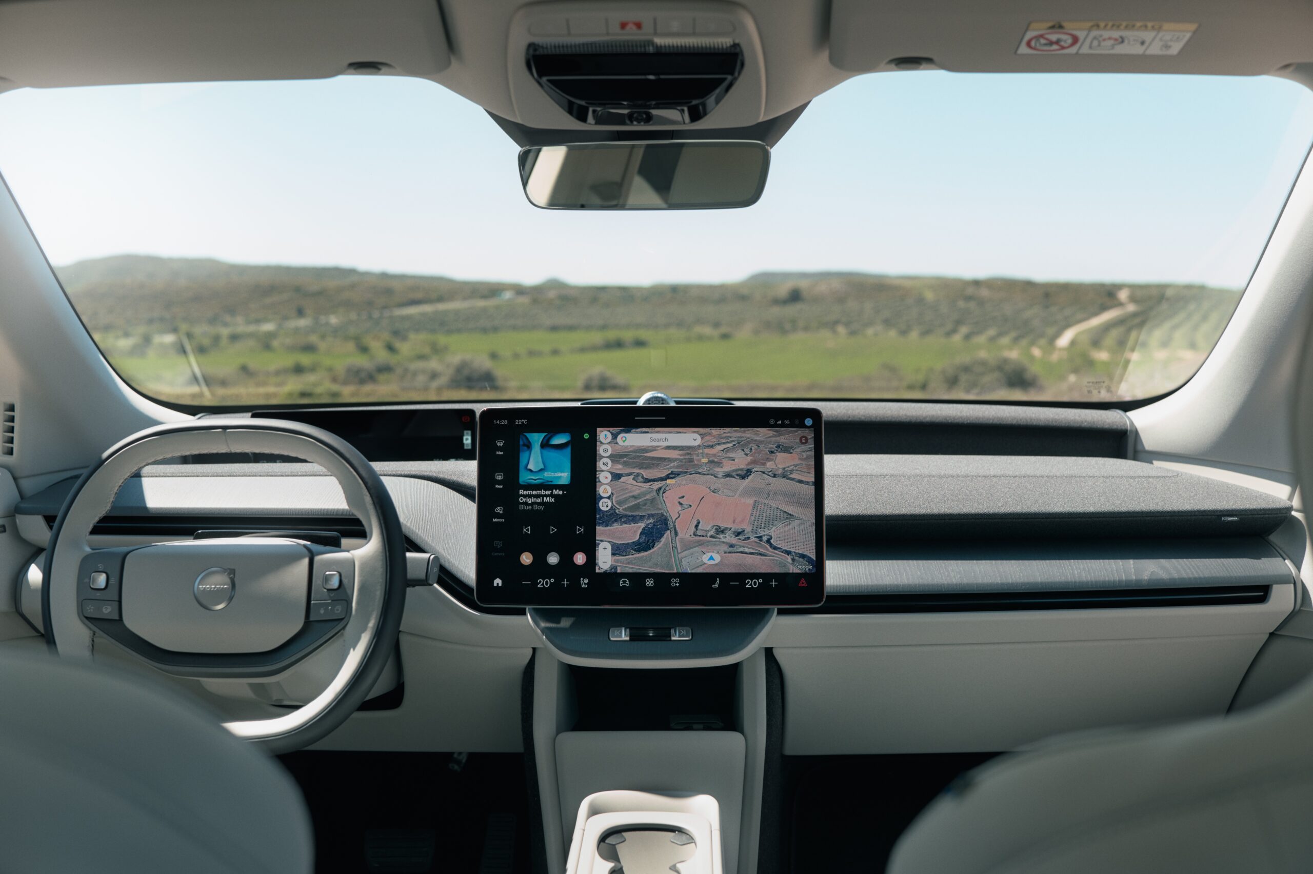 Volvo EX60 com Gemini não livra multa pelo CTB Interior de um carro moderno com volante cinza claro à esquerda e uma tela central exibindo um mapa e reprodutor de música. O para-brisa revela uma paisagem verde de colinas sob céu azul