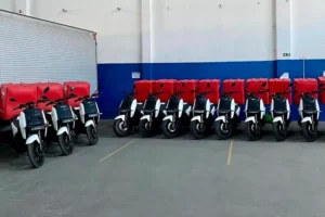 Nova linha de crédito amplia opções de trabalho em duas rodas Entregador com baú pilotando moto elétrica urbana em avenida brasileira, vista lateral em movimento, landscape