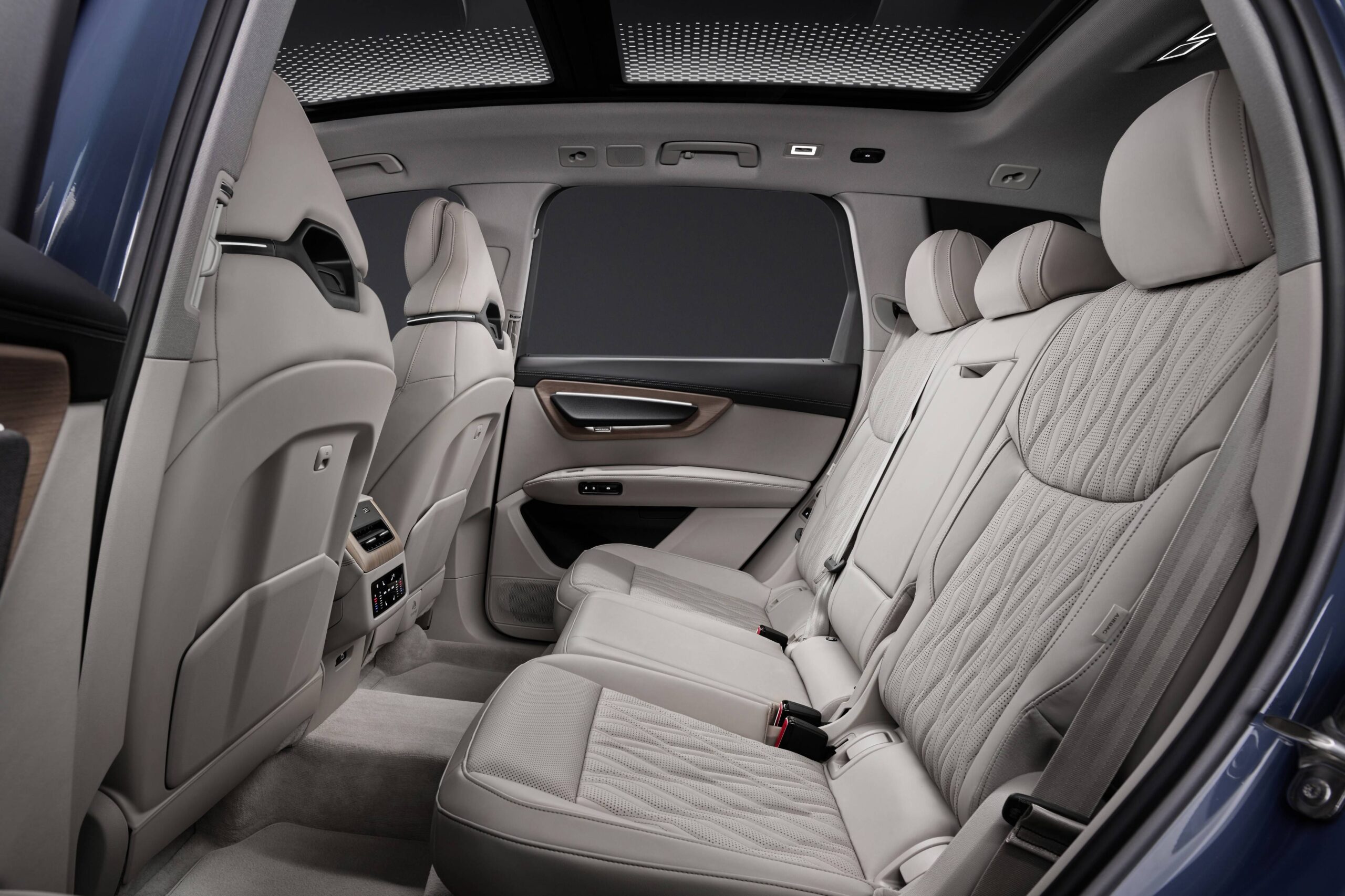 Novo Audi Q7 aposta em faróis divididos e luz no piso Interior de um carro moderno, mostrando os bancos traseiros em couro claro com detalhes acolchoados e um teto solar panorâmico. Há um apoio de braço central rebatido e controles de ventilação na parte de trás dos bancos dianteiros. As portas têm acabamento em madeira e metal, e o piso é acarpetado.