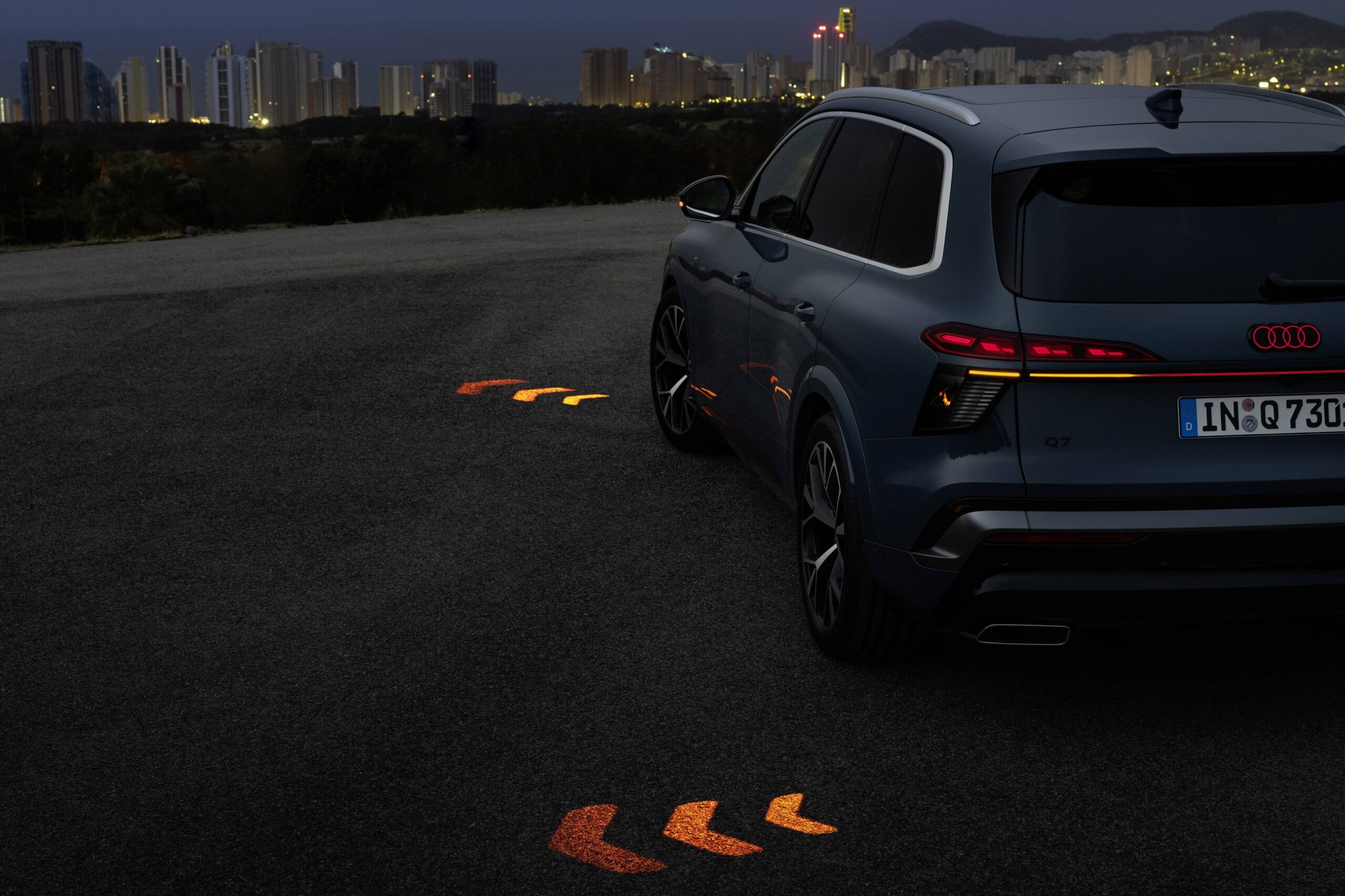 Novo Audi Q7 aposta em faróis divididos e luz no piso SUV Audi Q7 azul escuro estacionado em asfalto cinza escuro à noite, com setas luminosas laranja projetadas no chão. Ao fundo, uma cidade iluminada com prédios altos
