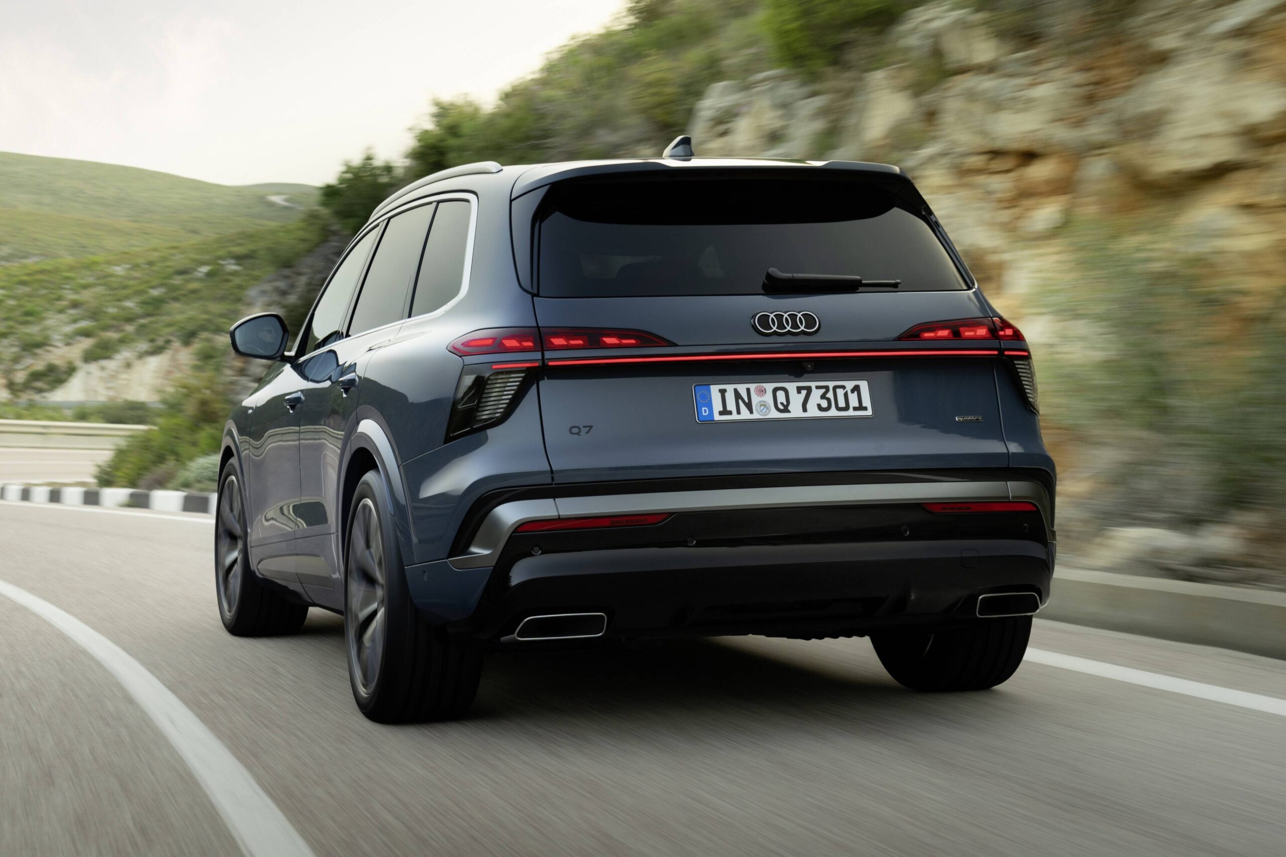 Novo Audi Q7 aposta em faróis divididos e luz no piso SUV Audi Q7 cinza-azulado visto por trás, em movimento numa estrada sinuosa com montanhas verdes ao fundo. As lanternas traseiras estão acesas, e a placa alemã IN Q 7301 é visível.