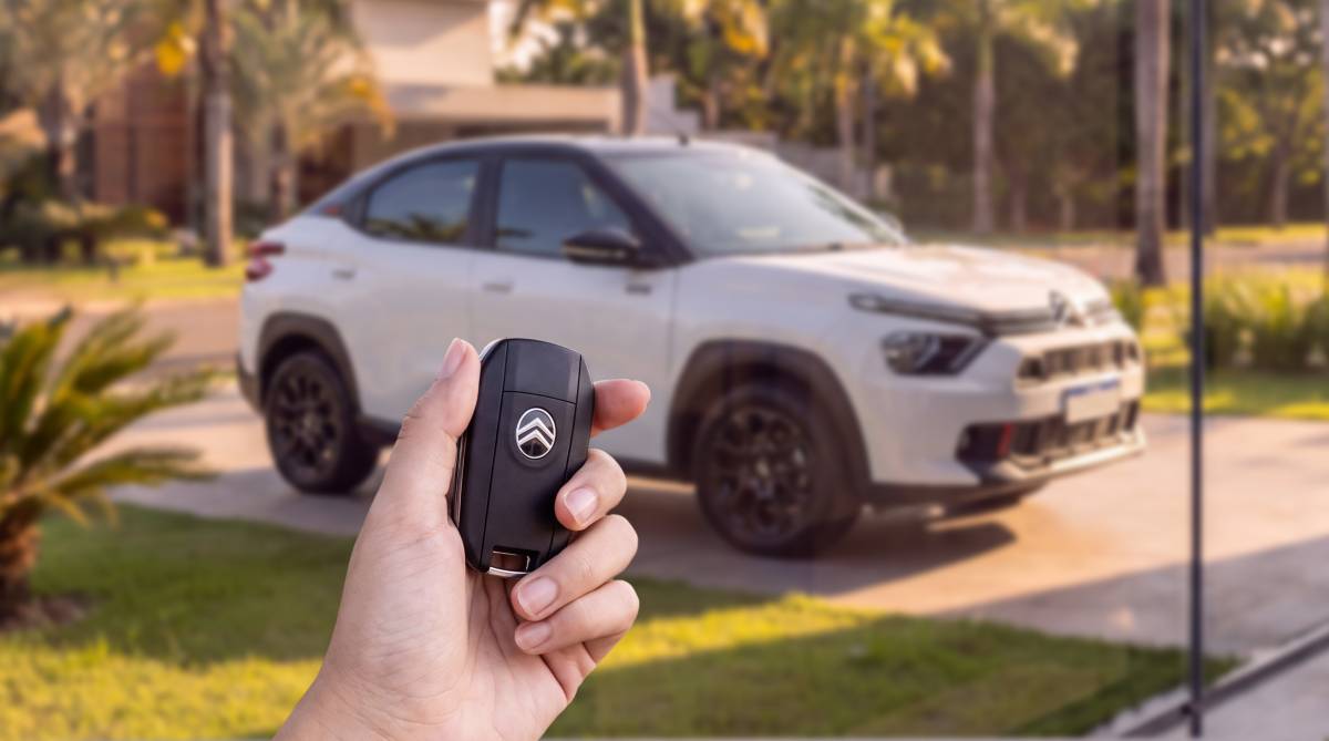 Citroën Basalt 2027: Preços partem de R$ 95.590 Mão segurando uma chave de carro preta com o logotipo da Citroën, com um carro branco e preto desfocado ao fundo, estacionado em uma garagem com gramado e palmeiras