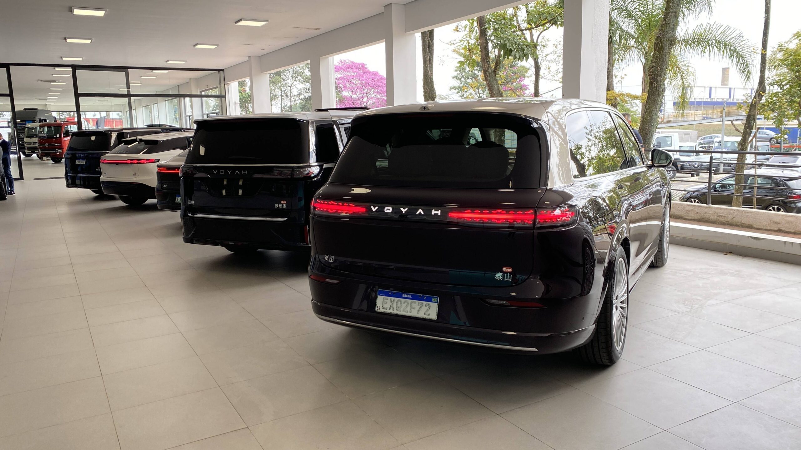Voyah no Brasil: Quatro modelos e até 1.530 km Vários carros de luxo, incluindo um SUV preto Voyah com placa brasileira, estacionados em uma concessionária com janelas grandes e árvores ao fundo