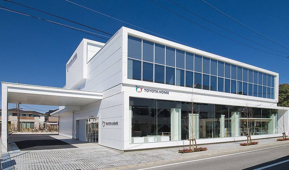 Toyota Home: Como funciona a casa modular de aço da Toyota Edifício moderno branco da Toyota Home com grandes janelas espelhadas, uma passarela coberta e estacionamento de paralelepípedos, sob céu azul claro
