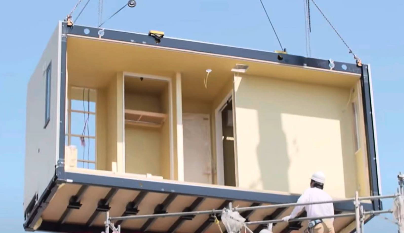 Toyota Home: Como funciona a casa modular de aço da Toyota Um módulo de casa pré-fabricada, com paredes internas claras e janelas, sendo içado por guindaste. Um trabalhador de capacete branco e camisa clara observa a operação de uma estrutura de andaimes abaixo