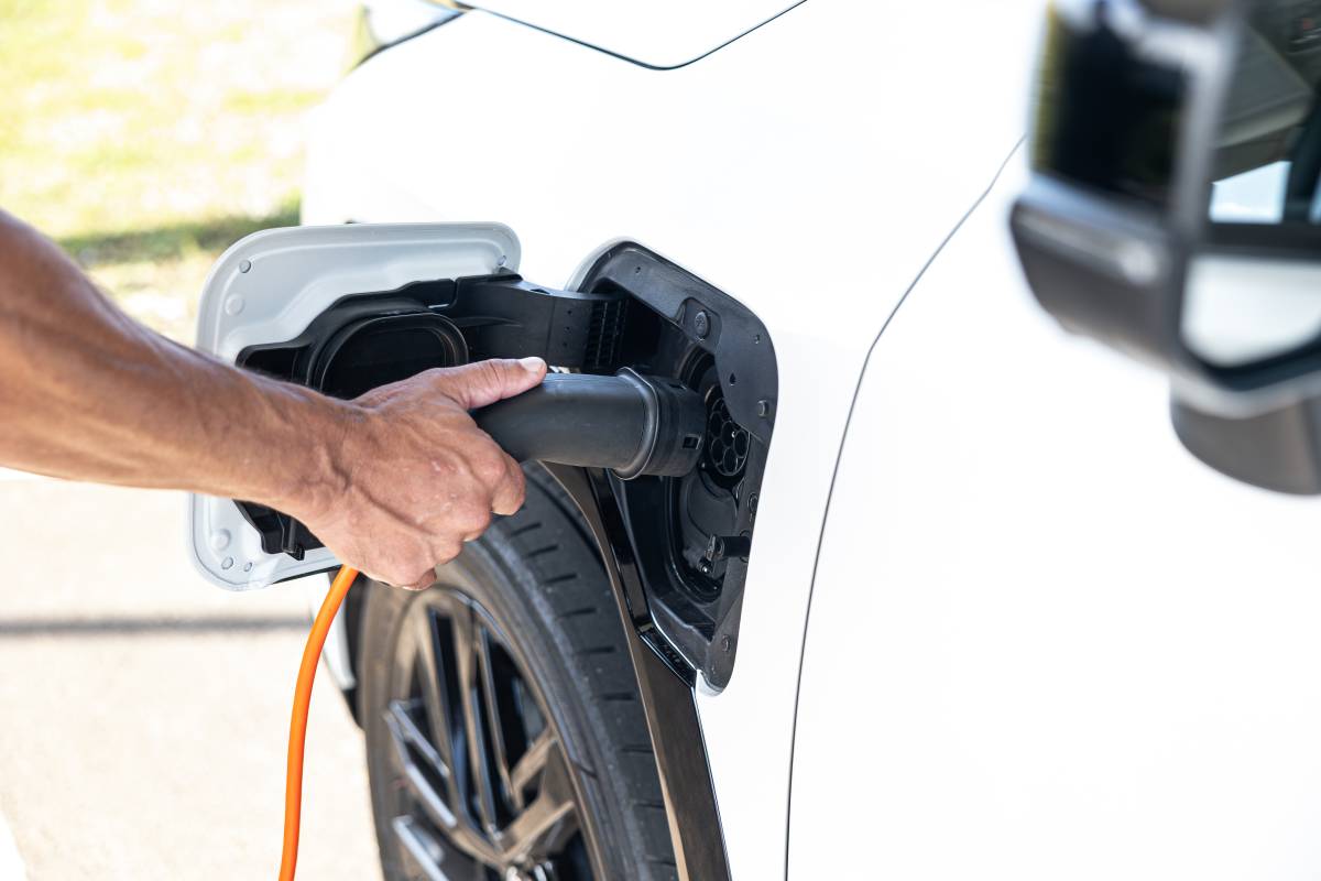 Toyota RAV4 PHEV: R$ 399.990 contra BYD Atto 8 Mão de uma pessoa conectando um cabo de carregamento preto e laranja na porta de um carro elétrico branco, com a roda dianteira visível