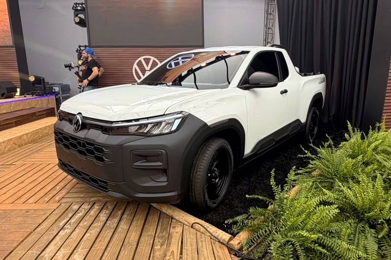 Volkswagen Tukan Robust mira briga com a Fiat Strada Endurance Picape branca com detalhes pretos, como para-choque e rodas, em um palco de madeira com plantas verdes ao lado. Ao fundo, um cinegrafista filma o veículo e o logo da Volkswagen aparece na parede