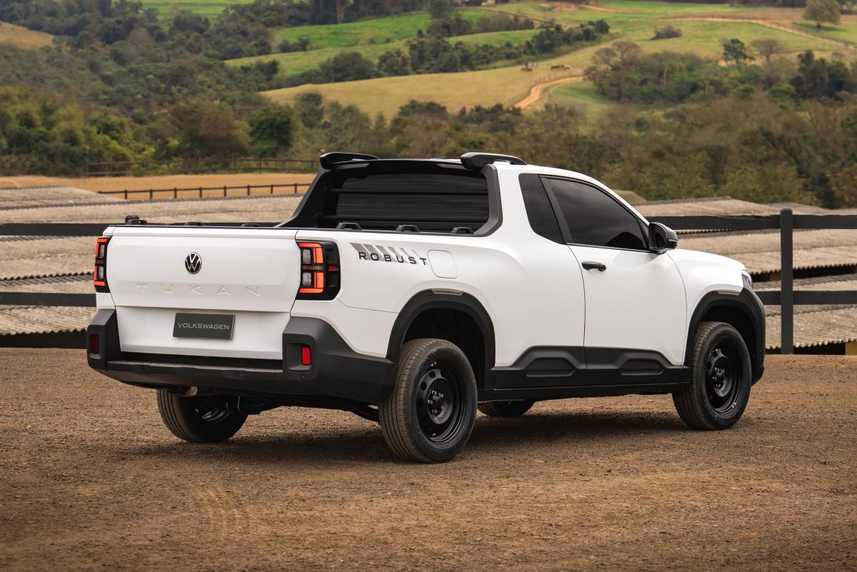 Volkswagen Tukan Robust mira briga com a Fiat Strada Endurance Picape branca Volkswagen Tukan Robust, vista de traseira e lateral, estacionada em terreno de terra batida. Possui rodas pretas, detalhes em plástico preto na parte inferior da carroceria e na caçamba, e lanternas traseiras verticais com iluminação em LED. Ao fundo, uma cerca de madeira e colinas verdes com vegetação densa