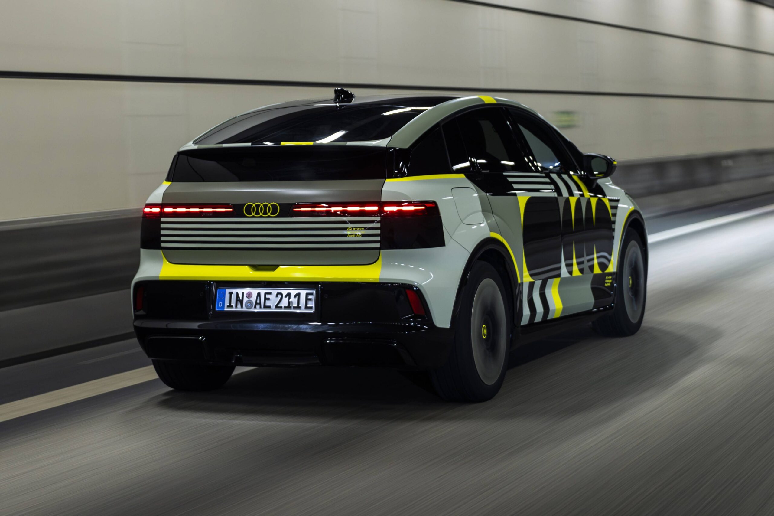 Audi A2 2026 existe? Rumor elétrico mira 476 km Carro Audi camuflado em cinza, preto e amarelo neon, visto por trás e lateralmente, em movimento rápido dentro de um túnel iluminado