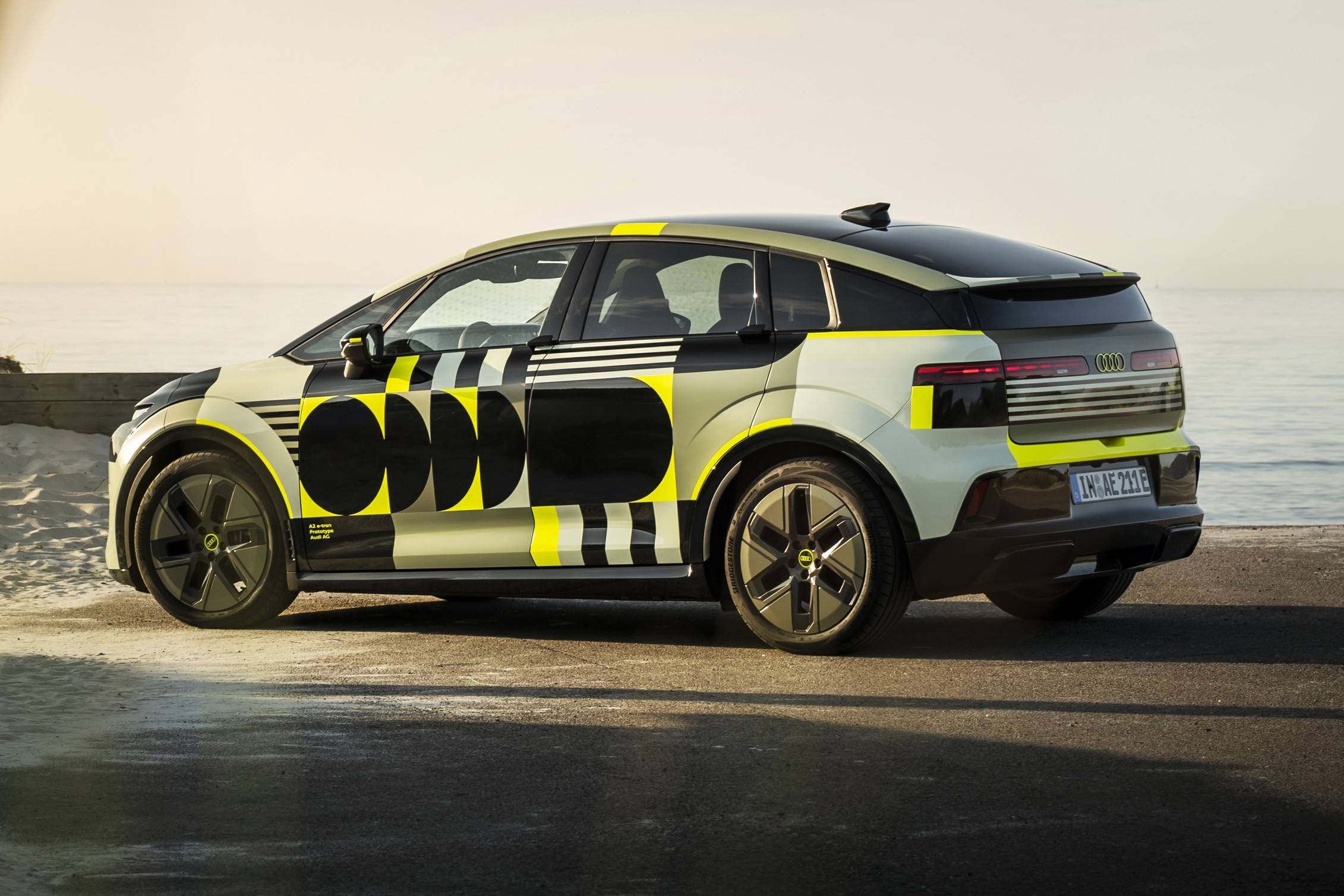 Audi A2 2026 existe? Rumor elétrico mira 476 km Carro elétrico camuflado com padrões geométricos em preto, amarelo e cinza claro, estacionado em uma estrada de asfalto perto do mar, sob luz solar