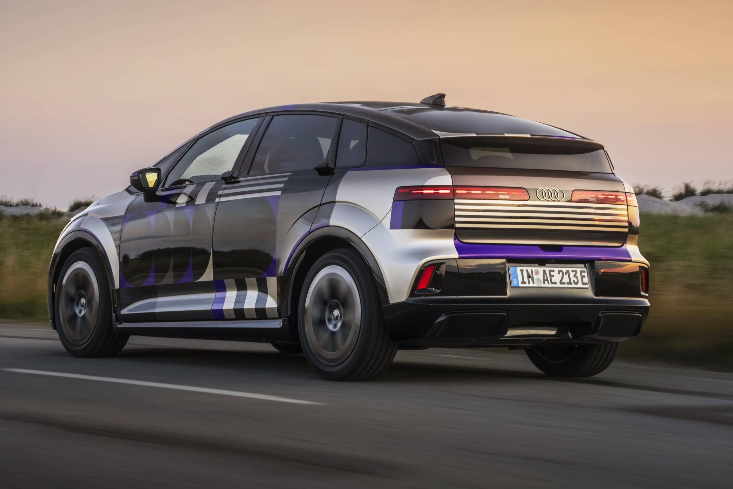 Audi A2 2026 existe? Rumor elétrico mira 476 km Carro Audi elétrico camuflado com padrões geométricos em preto, prata e roxo, visto de traseira lateral, em movimento numa estrada, com céu claro ao fundo