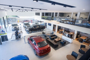 Fachada horizontal de concessionária brasileira movimentada, com compradores analisando carros novos e vendedores atendendo clientes, landscape